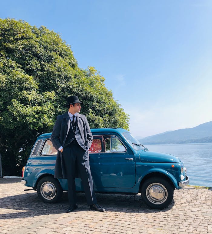 Man in gray suit and hat leaning on blue vintage car by a lake in Ghiffa, Italy.