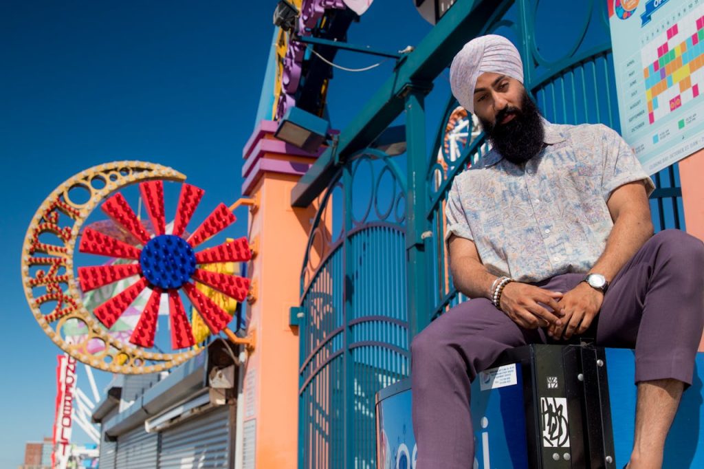 pexels-photo-2890694 A fashionable man in a turban poses at a colorful amusement park under clear skies.